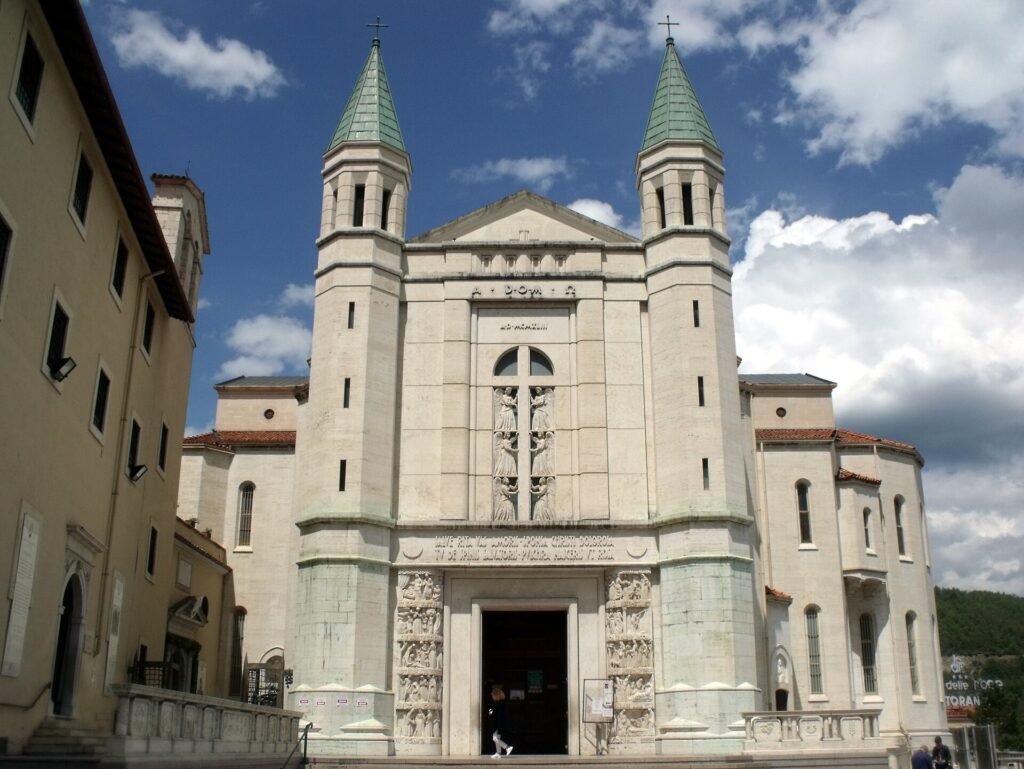 The pale stone facade of the Basilica of Santa Rita in Cascia, with twin green-capped bell towers under a cloudy blue sky