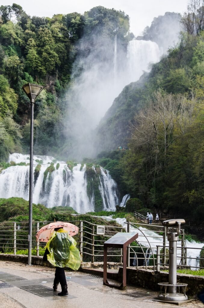 Un visitatore con poncho verde e ombrello al belvedere inferiore con ringhiera, la cascata e i suoi spruzzi sullo sfondo