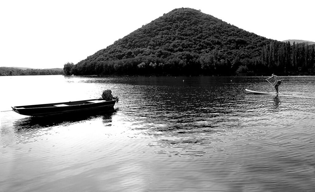 Foto in bianco e nero di una piccola barca e di un pagaiatore sull’acqua ferma sotto una collina boscosa a forma di cono