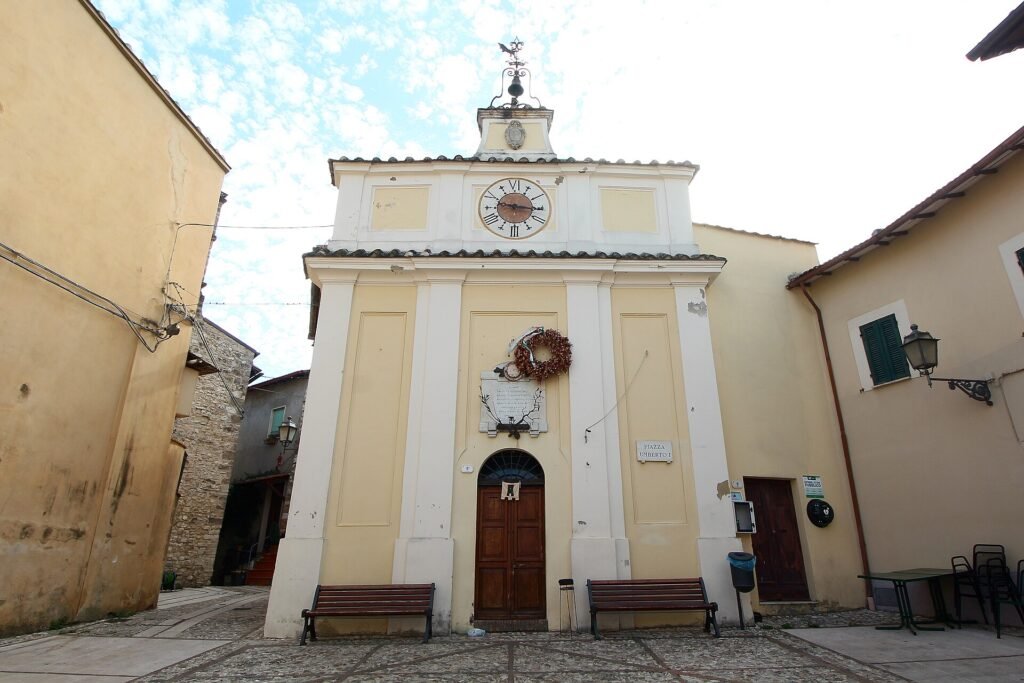 Edificio giallo chiaro con lesene bianche, un quadrante d’orologio sul fastigio e una piccola campana in cima