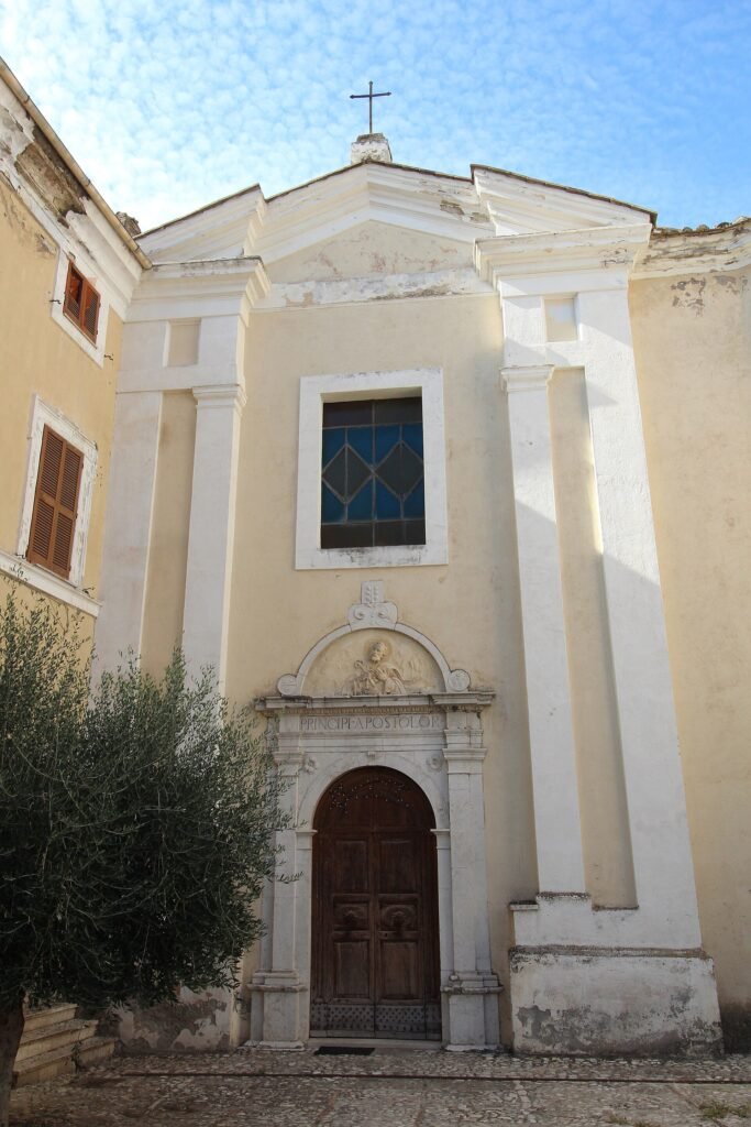 Facciata gialla chiara con lesene bianche, una finestra quadrata e un portale in pietra scolpita, con un olivo davanti