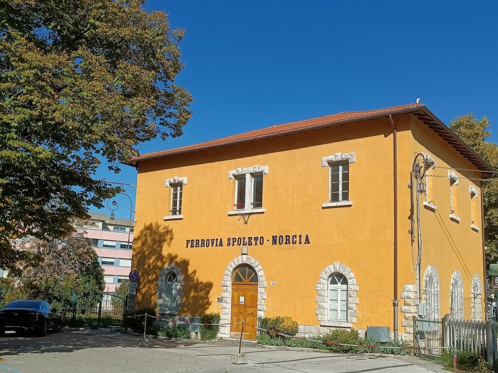 Un edificio giallo a due piani con la scritta Ferrovia Spoleto-Norcia sulla facciata, sotto un cielo azzurro