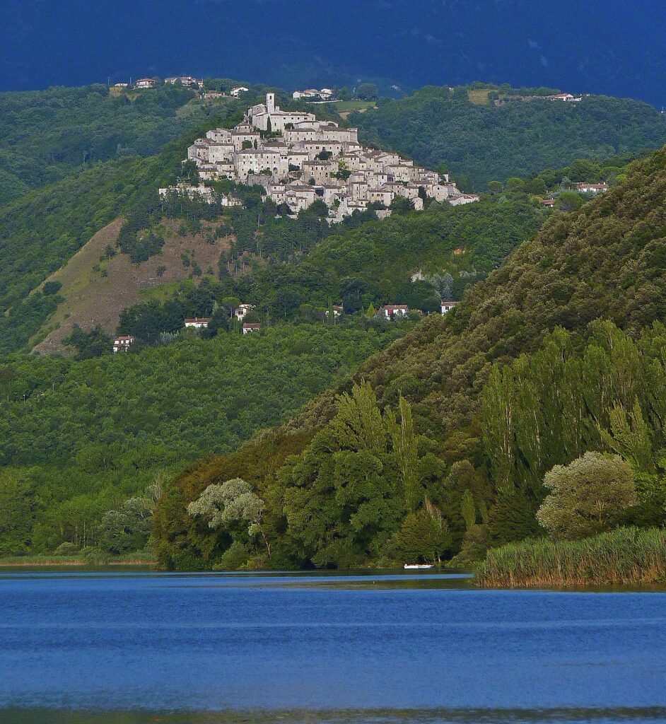 Le case chiare di pietra di Labro in cima a una collina verde sopra i boschi, con canne e lago azzurro in primo piano