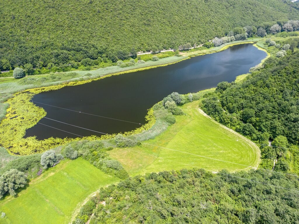 Veduta aerea del piccolo e scuro Lago di Ventina, circondato da canneti e prati verdi, con colline boscose dietro