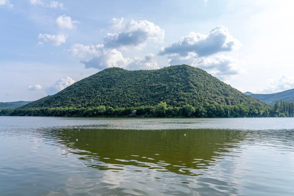Una collina boscosa e tondeggiante sopra il Lago di Piediluco, riflessa nell’acqua calma sotto un cielo azzurro con nuvole