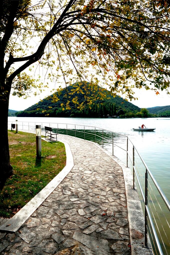 Un vialetto lastricato con ringhiera lungo il lago sotto un albero, una collina boscosa a cono oltre l’acqua calma