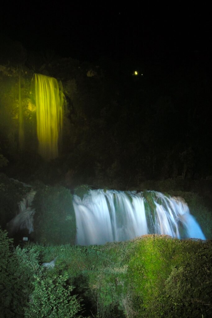 La cascata di notte, il salto superiore illuminato di giallo-verde nel buio e quello inferiore di bianco