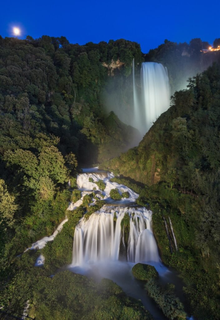 Marmore Falls floodlit at night, the upper leap and lower cascades glowing white against dark woods and a deep blue sky