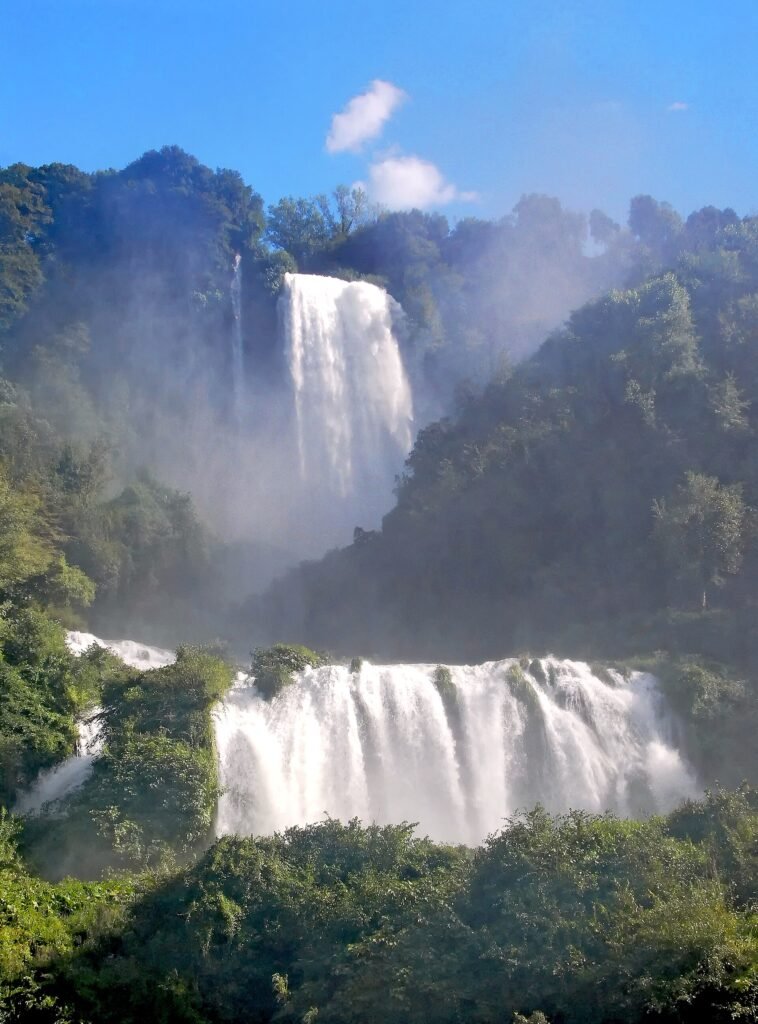 Il salto superiore della Cascata delle Marmore tra i boschi e l'ampia cascata inferiore sotto, con il cielo azzurro