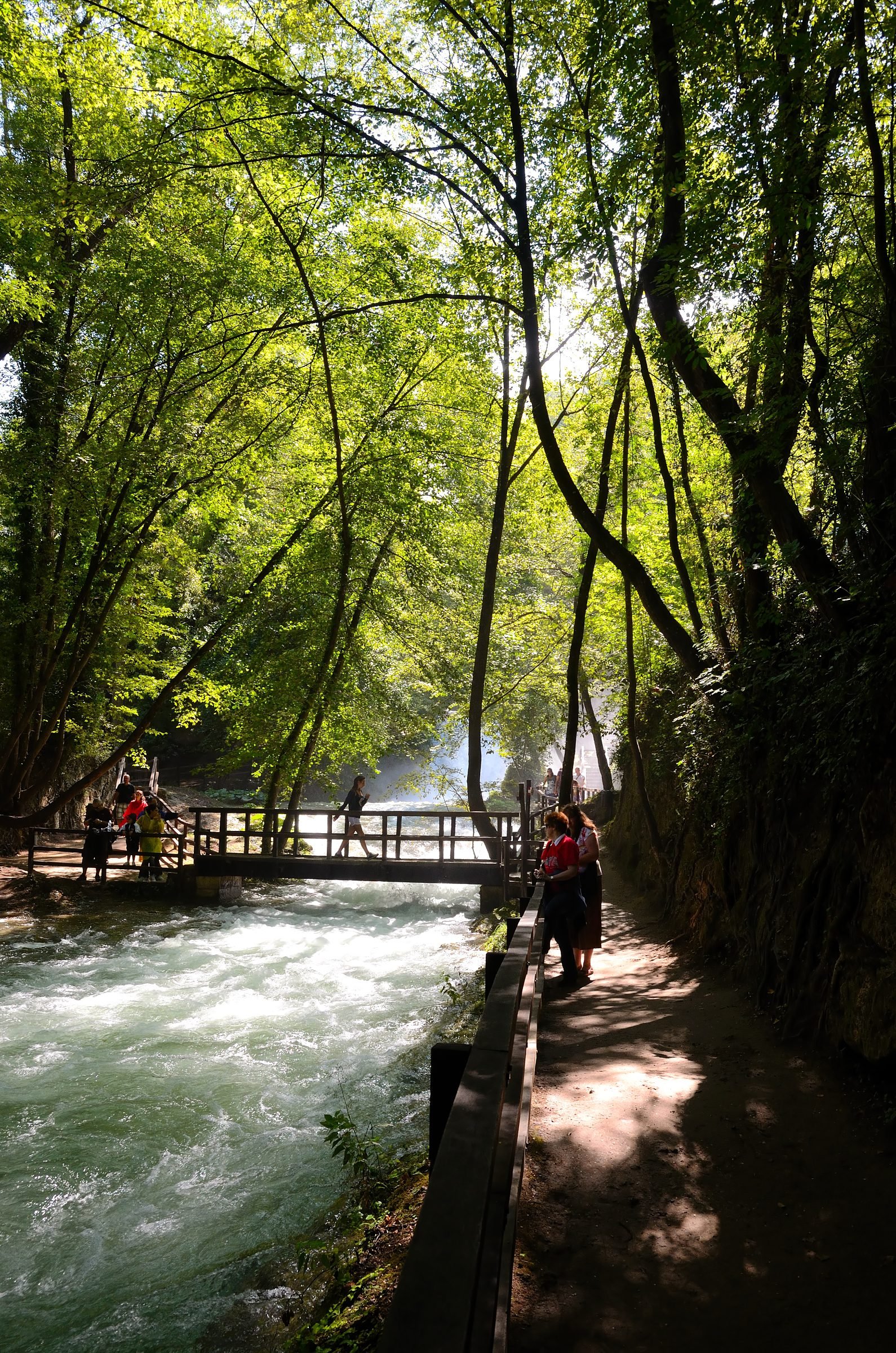 A gravel riverside path in the Marmore Falls park with a wooden footbridge and walkers