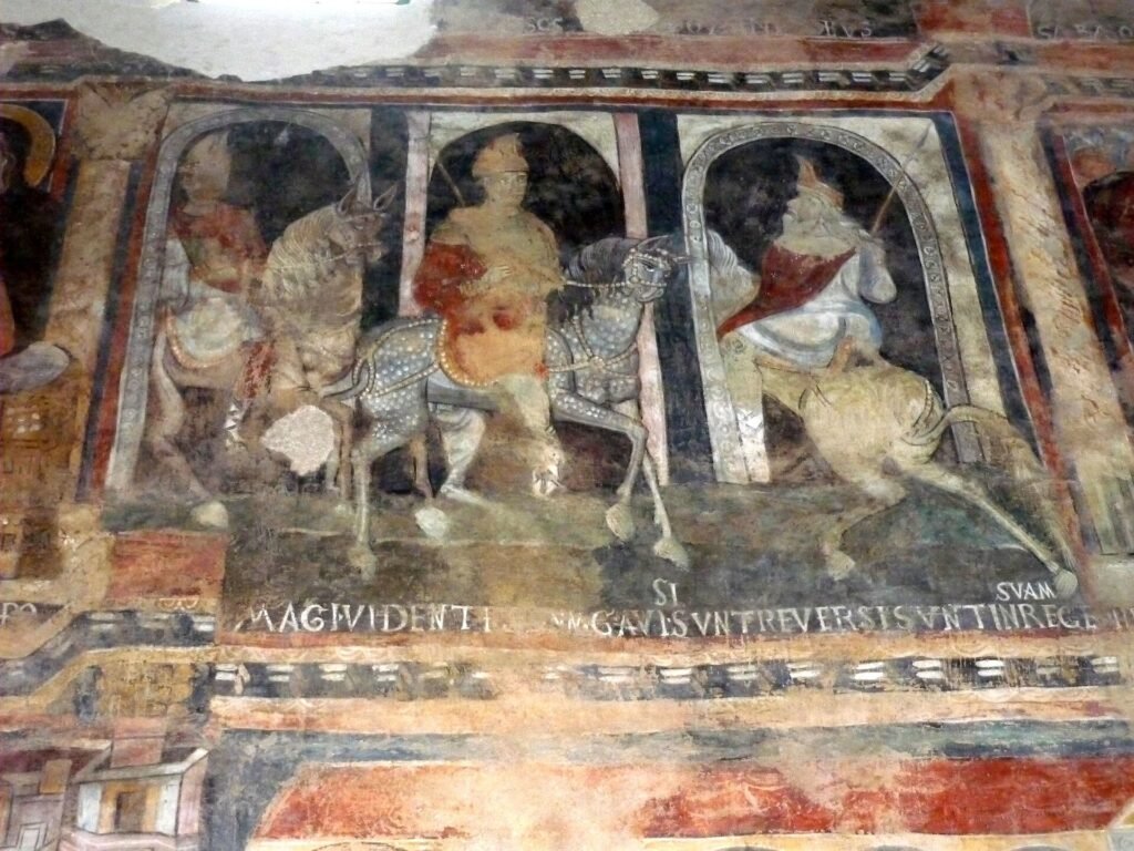Faded medieval fresco of three crowned riders on horseback under painted arches, with a Latin inscription below