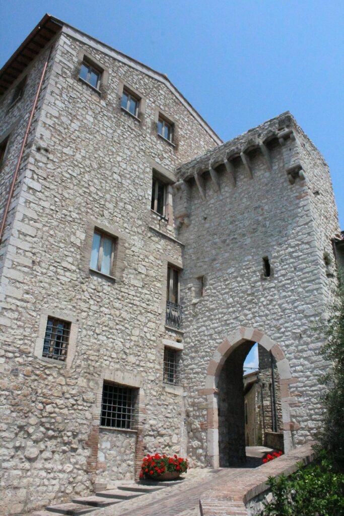 Alta casa-torre in pietra con un arco di passaggio alla base, gradini e fiori rossi davanti