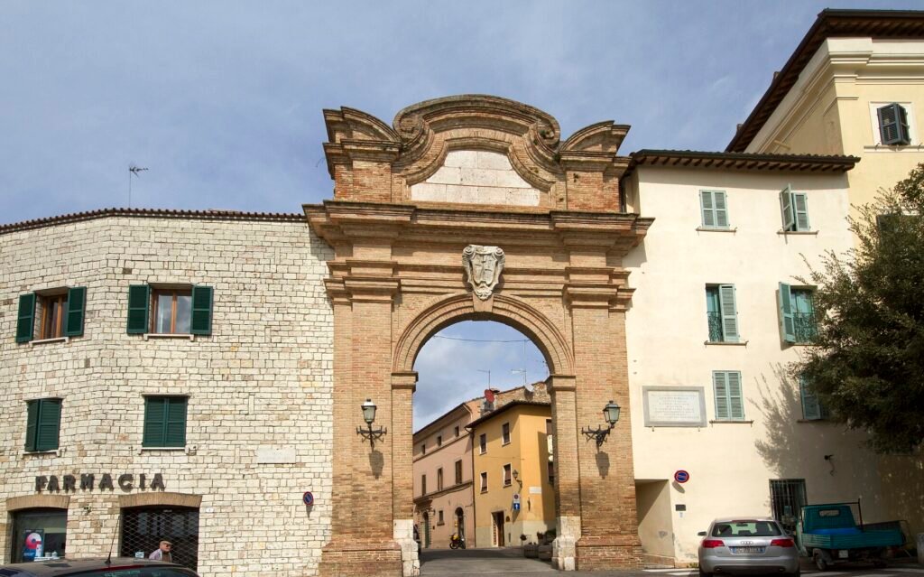 Un portale ad arco in mattoni e pietra tra edifici chiari a San Gemini, con l’insegna di una farmacia a sinistra