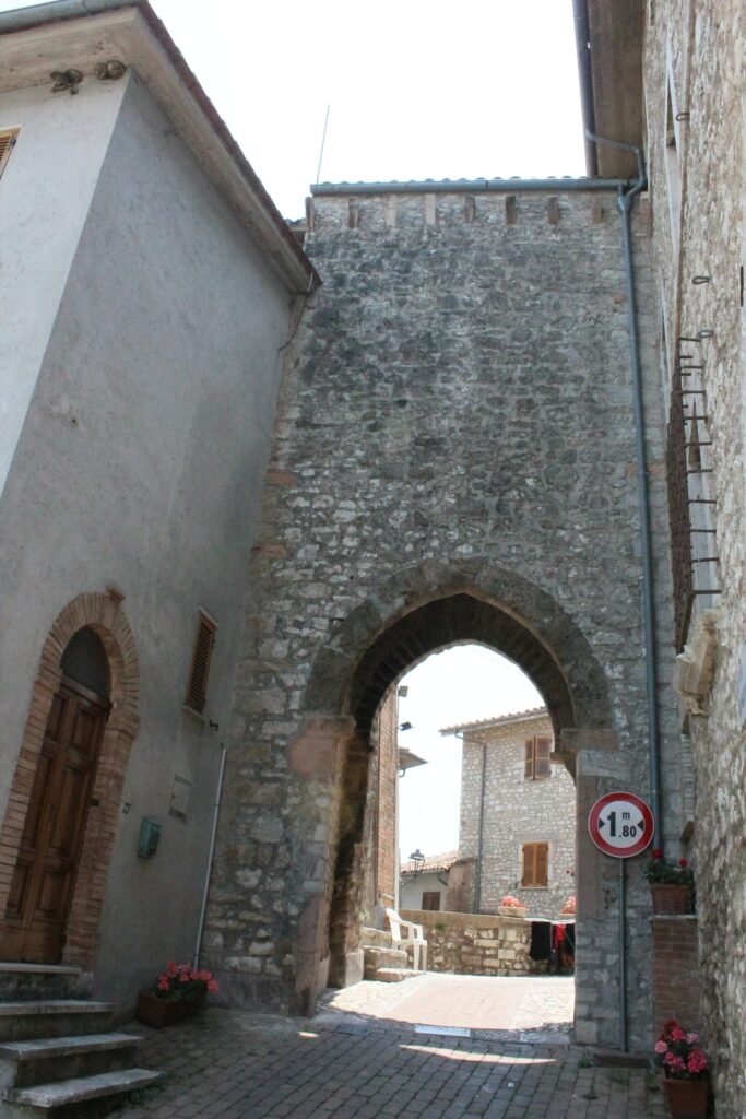Porta ogivale in pietra tra due case, con un vicolo stretto e vasi di fiori oltre l’arco