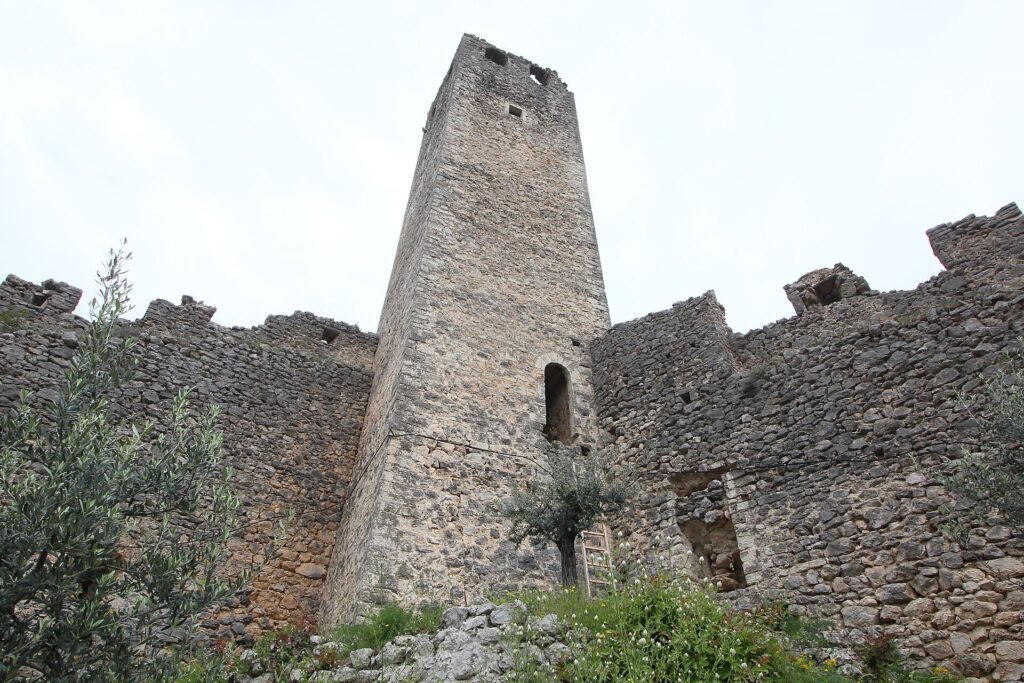 Un'alta torre quadrata in pietra dietro mura medievali diroccate, con ulivi ed erba in primo piano