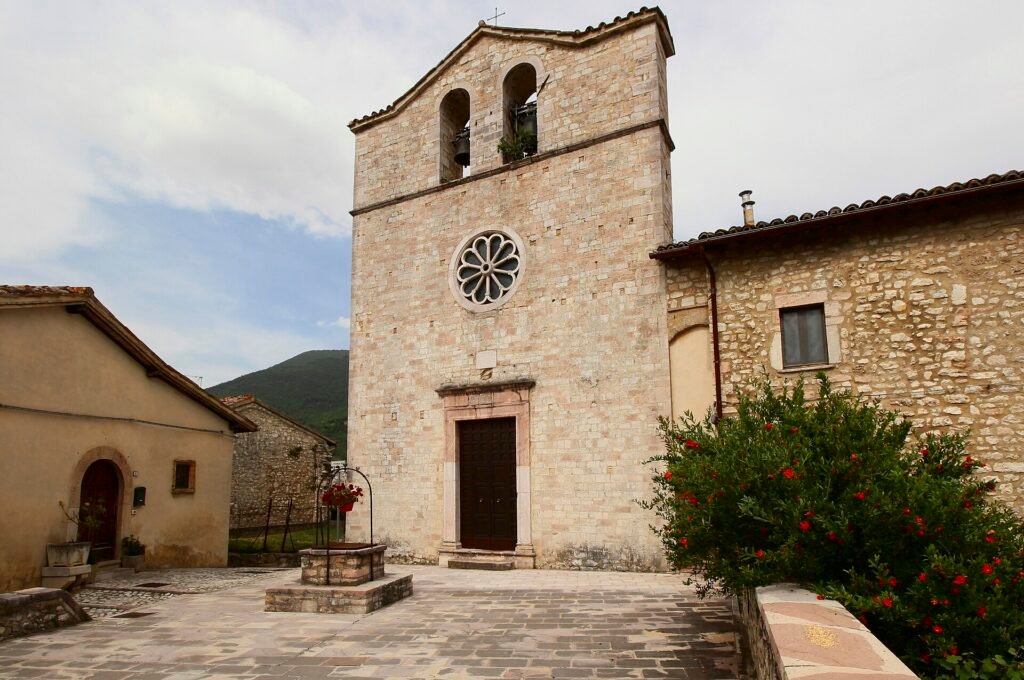 Semplice facciata in pietra con rosone e campanile a vela, su una piazzetta lastricata con un pozzo