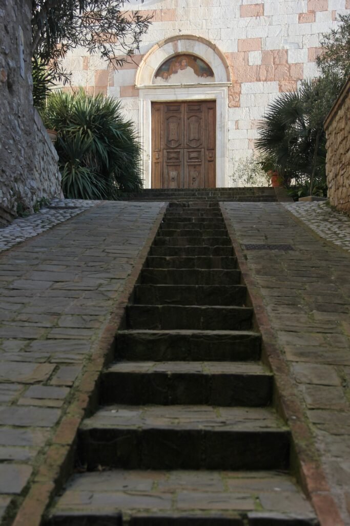 Ampi gradini di pietra che salgono alla porta in legno di una chiesetta con facciata in pietra rosa e bianca
