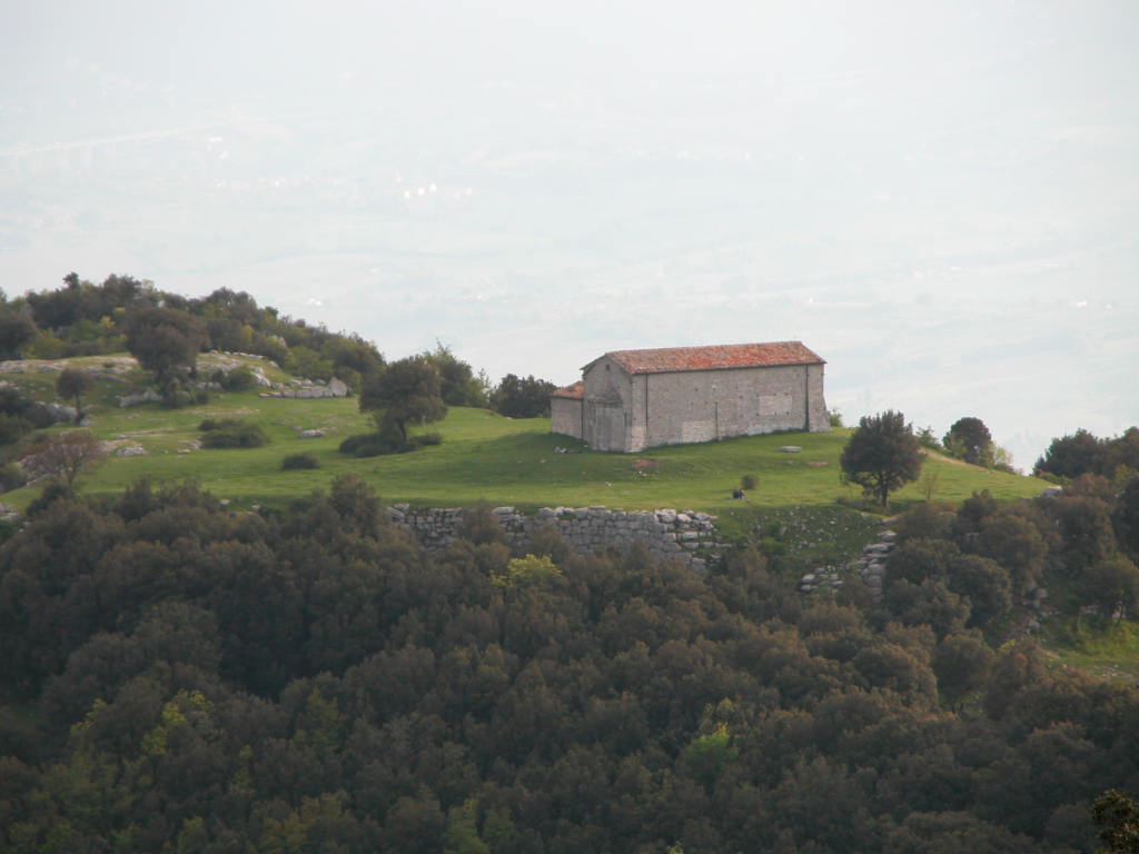 Una piccola chiesa di pietra con tetto in coppi su un pianoro erboso, con antiche mura di pietra sotto e bosco davanti