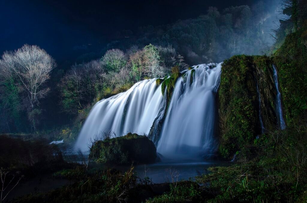 A long-exposure night view of a wide lower leap of Marmore Falls, smooth white water between dark mossy rocks and bare trees