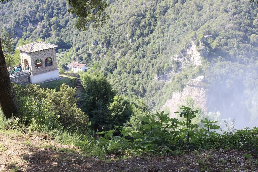 Piccolo padiglione panoramico in pietra con archi su uno sperone boscoso, visitatori alla ringhiera e spruzzi accanto