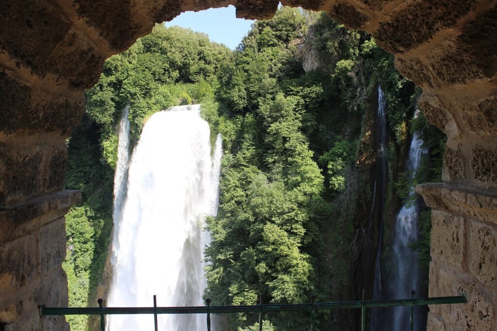 Vista attraverso un arco di pietra sul primo salto bianco della Cascata delle Marmore tra pareti boscose